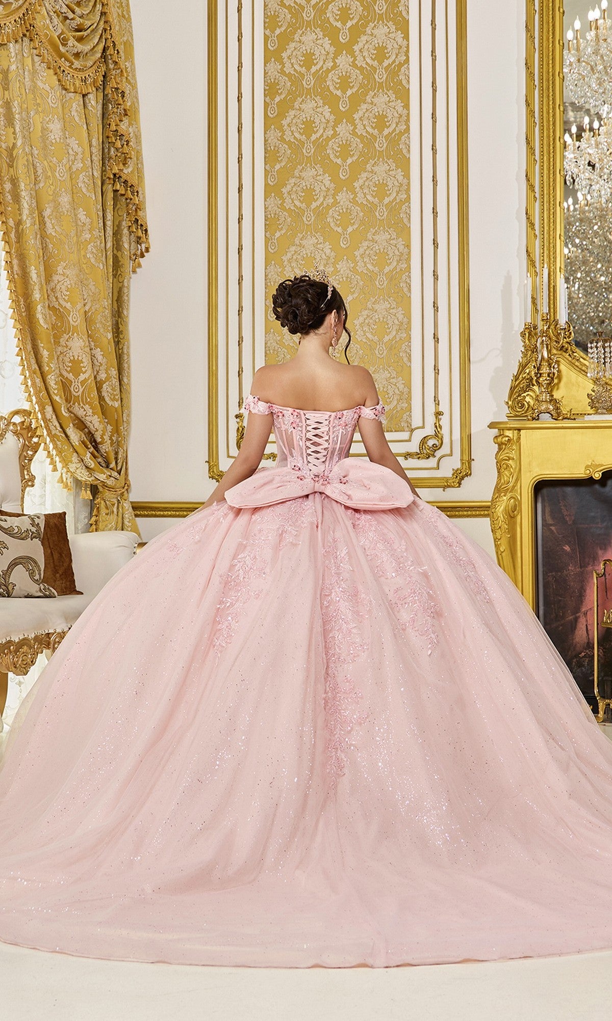 A woman in the Quinceanera Dress 5090 By Dancing Queen, a blush pink off-the-shoulder ball gown with glitter tulle layers, stands facing away in an ornate gold and white room.